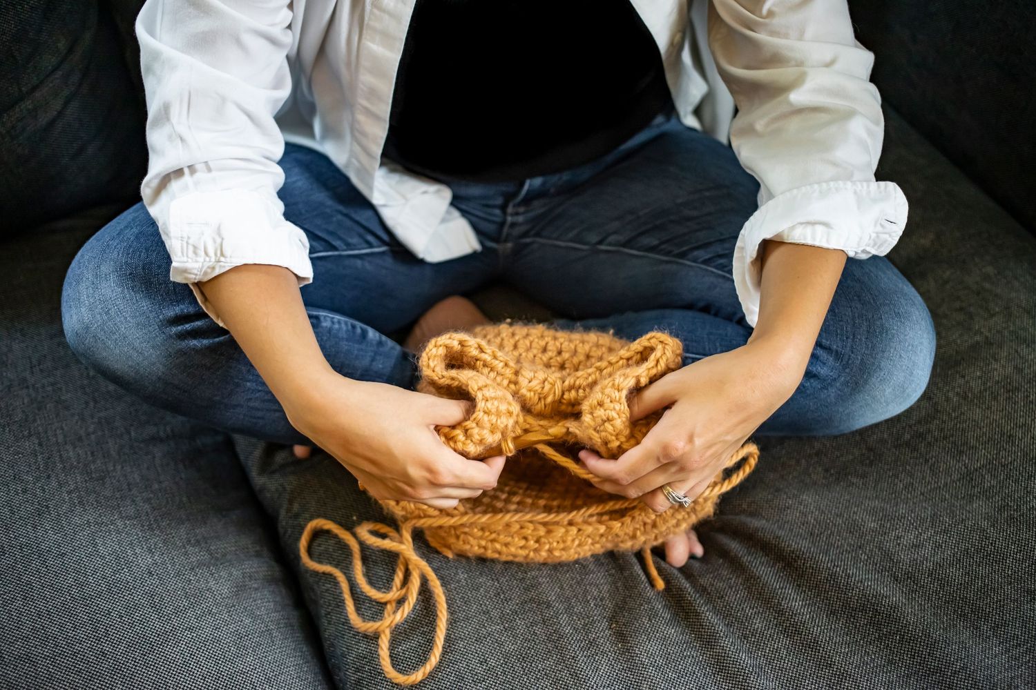 Person knitting with caramel colored yarn while sitting cross-legged on dark couch.