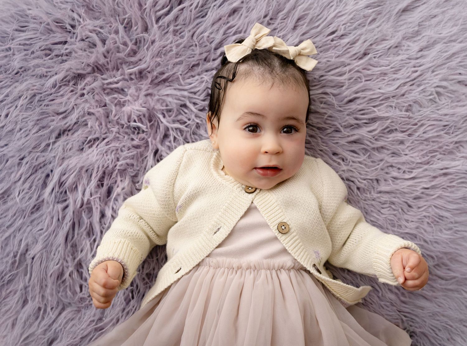 Toddler baby girl with pink pigtail bows laying on a lavender fur and looking up at the camera
