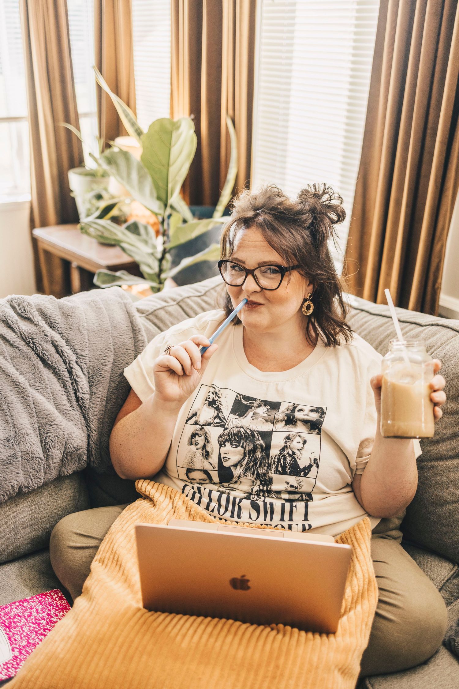 Woman in Taylor Swift shirt sipping iced coffee while working on MacBook on couch.