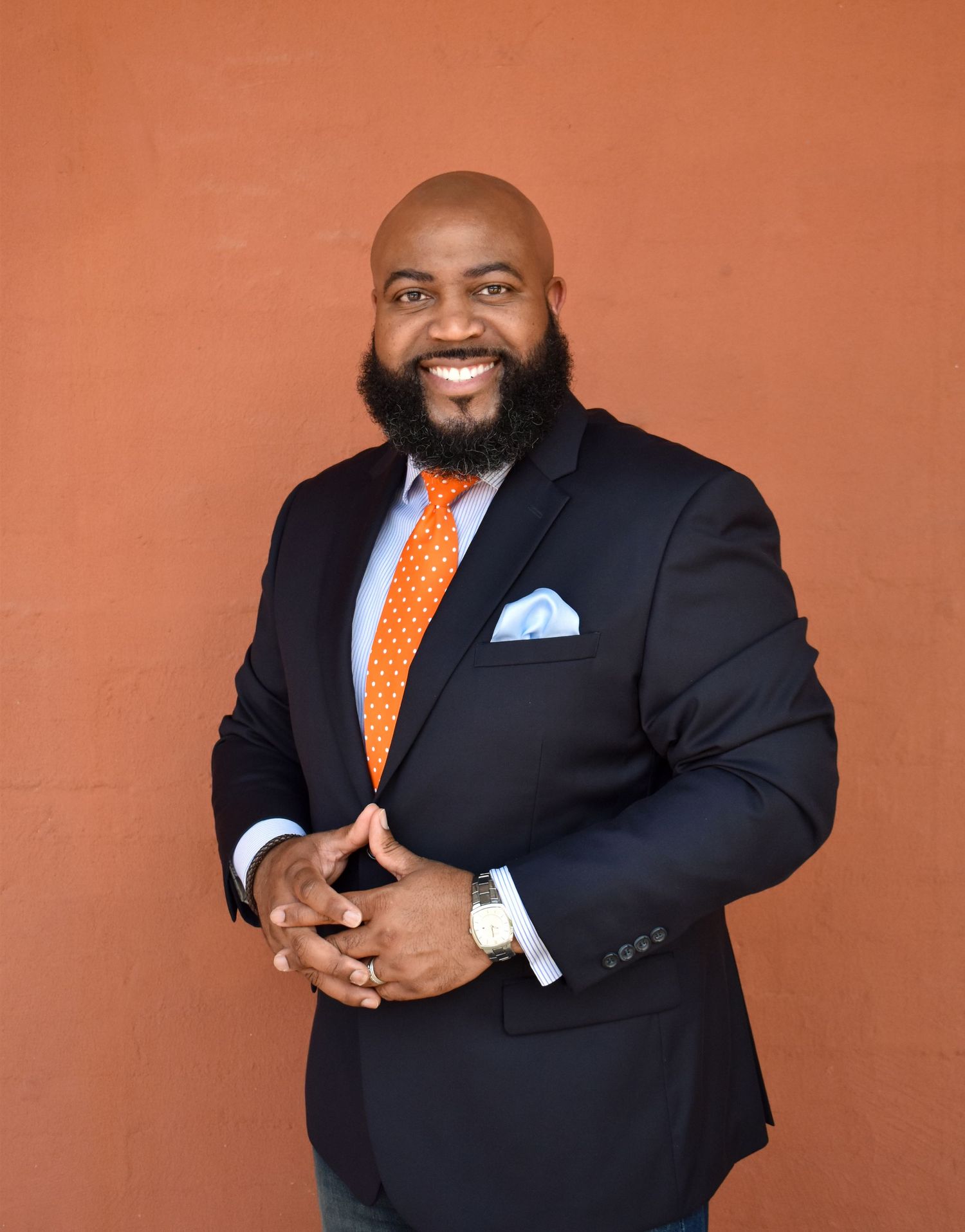 Professional man in navy suit and orange tie with pocket square smiles warmly against orange backdrop.