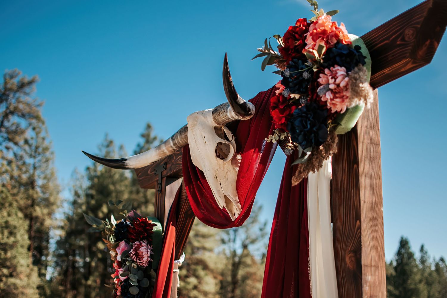 Wedding arch decorated with red ribbons, floral arrangements and a cow skull against a bright blue sky in Oregon.