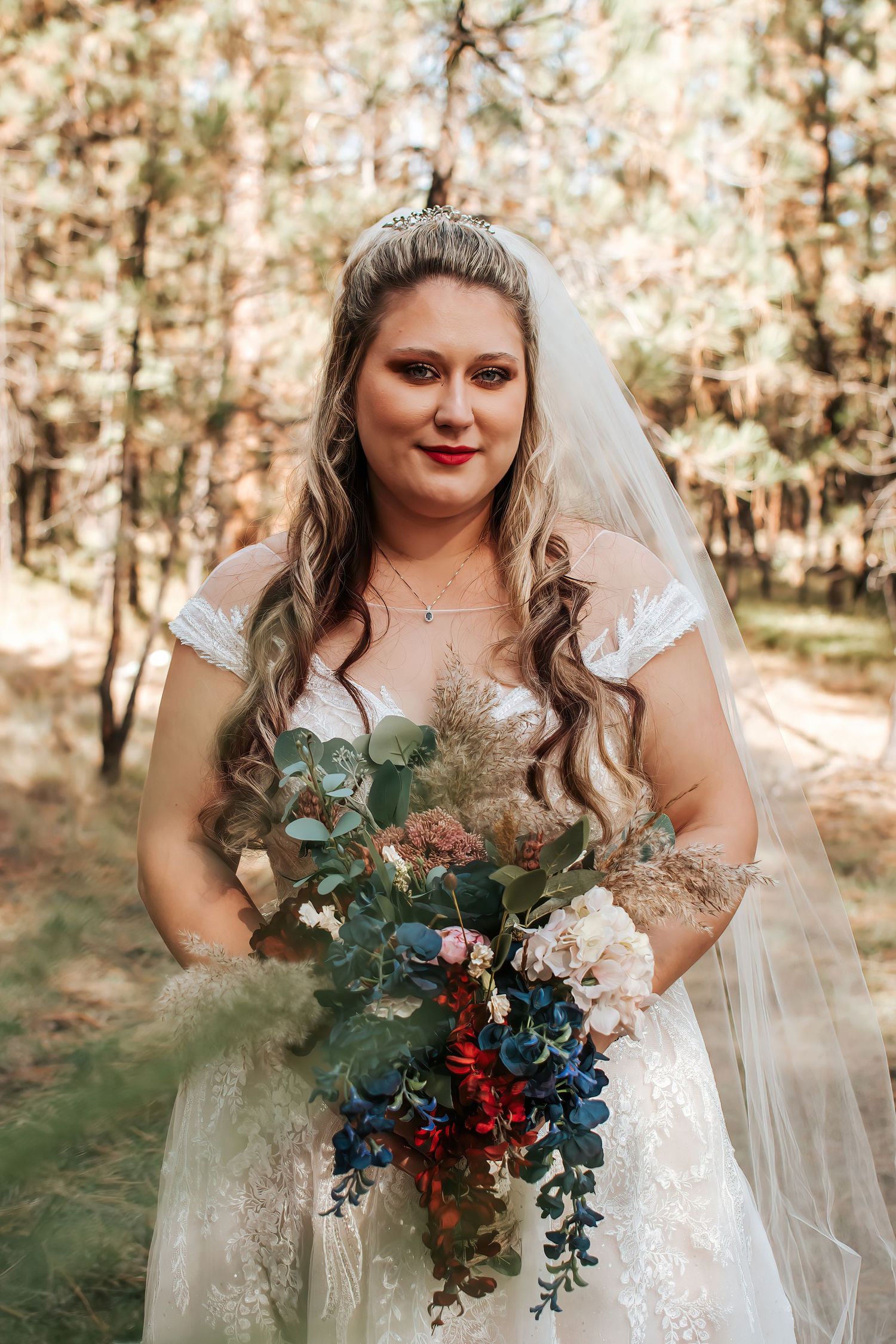 Bride holding a vibrant bouquet of blue and red flowers with eucalyptus in a forest setting in Ukiah, Oregon.