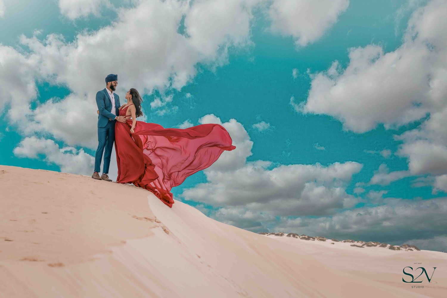 A couple stands atop a sand dune with a flowing red dress against a vibrant turquoise sky.