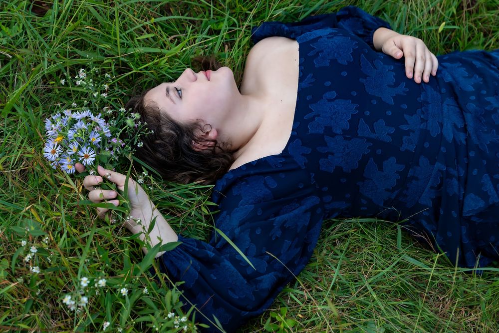 A person in a navy blue dress lies in lush green grass surrounded by delicate white wildflowers. Senior portrait Morgantown wv