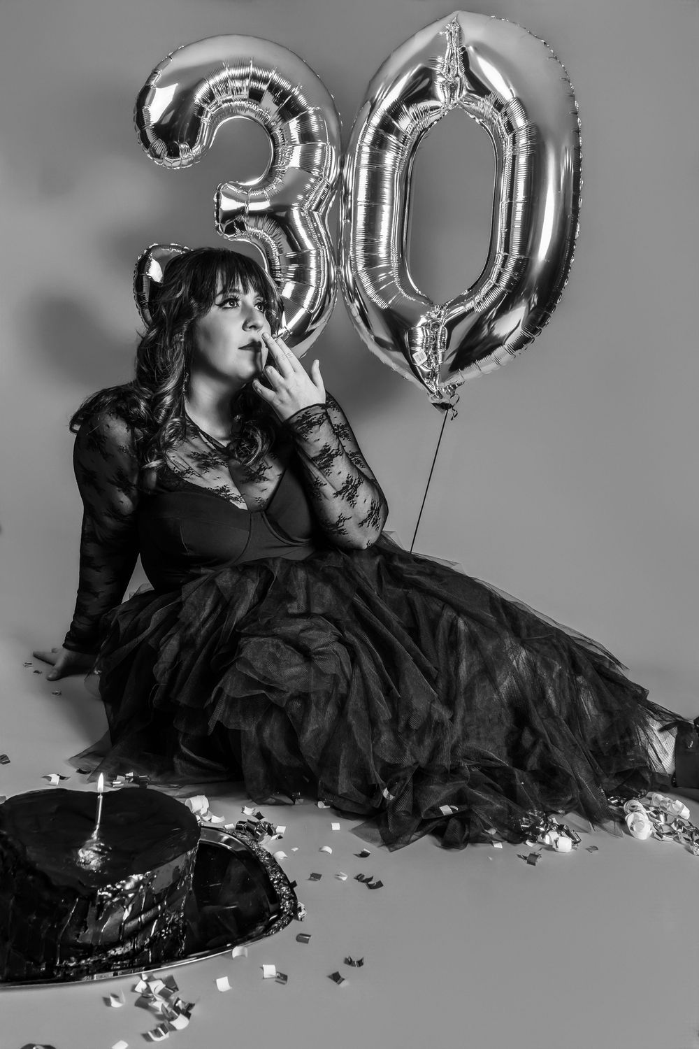 Woman in black lace gown sits beside smashed cake and silver 30 birthday balloons in dramatic black and white photo.