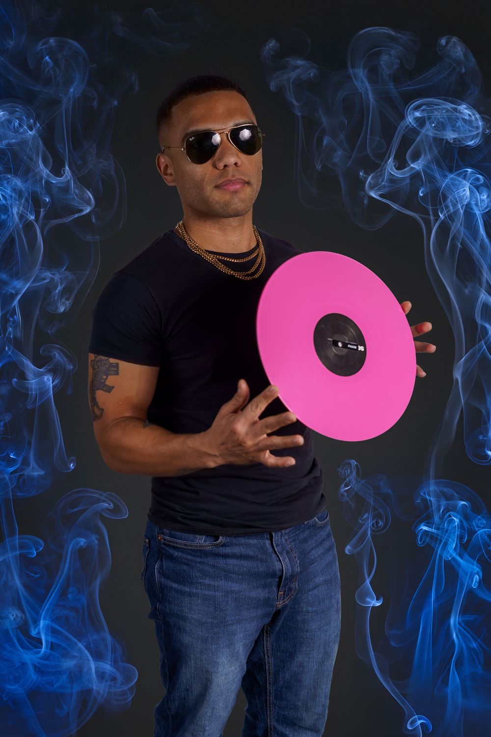 DJ in sunglasses and gold chains holds a pink vinyl record, surrounded by blue smoke on a dark background.