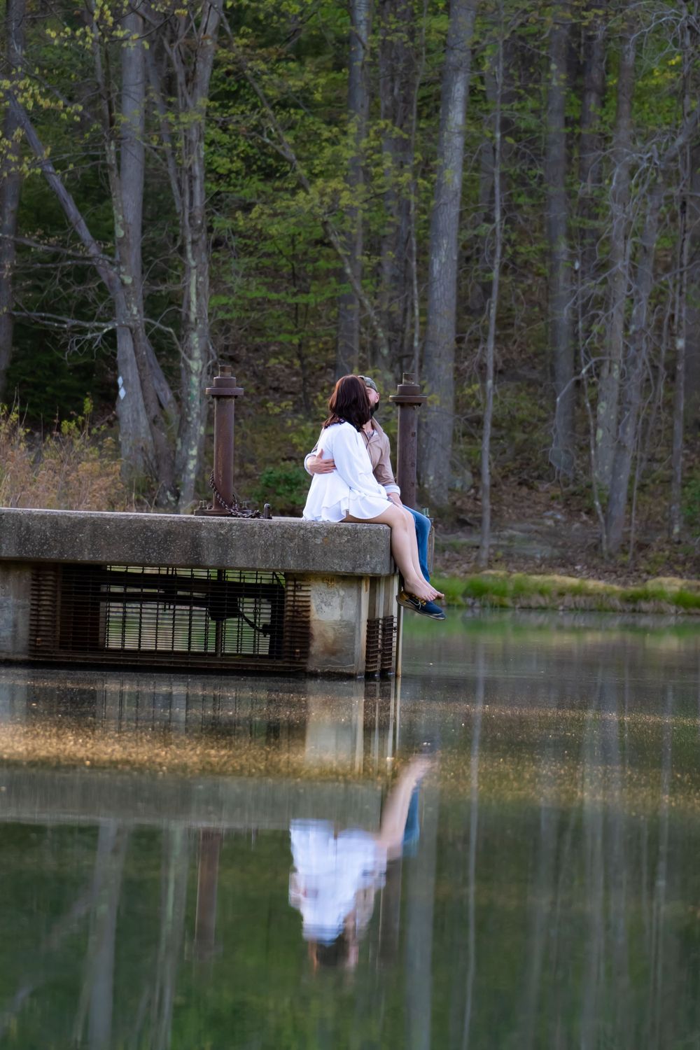 Reflective scene of someone sitting on concrete structure above calm lake water.