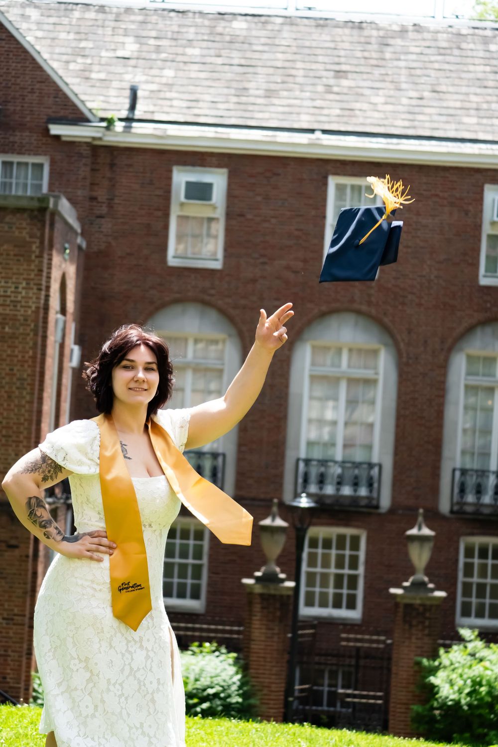 Graduate in white dress and gold stole throws cap in celebration outside brick university building.