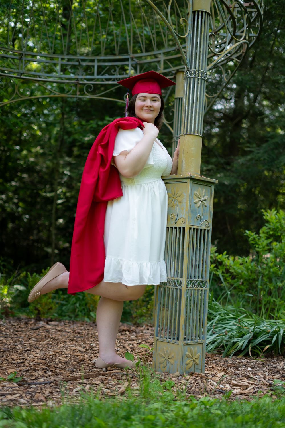Graduate in red cap and white dress poses elegantly by garden architecture.
