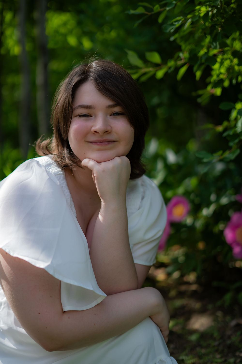 Person in white dress poses outdoors against lush green garden background.