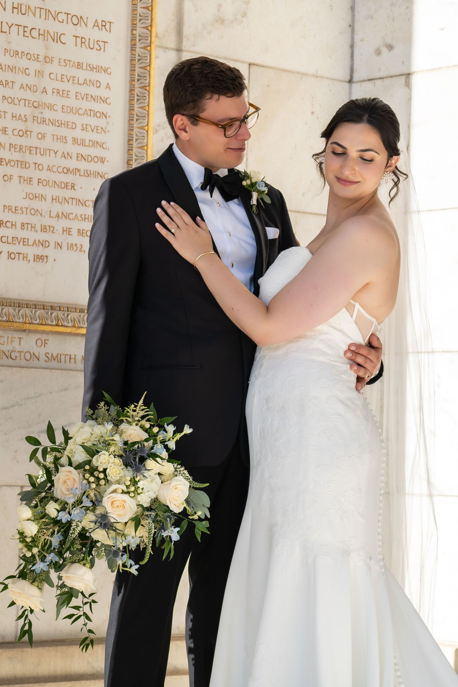 Elegant wedding couple in formal attire dancing together near a white marble wall with floral decorations.