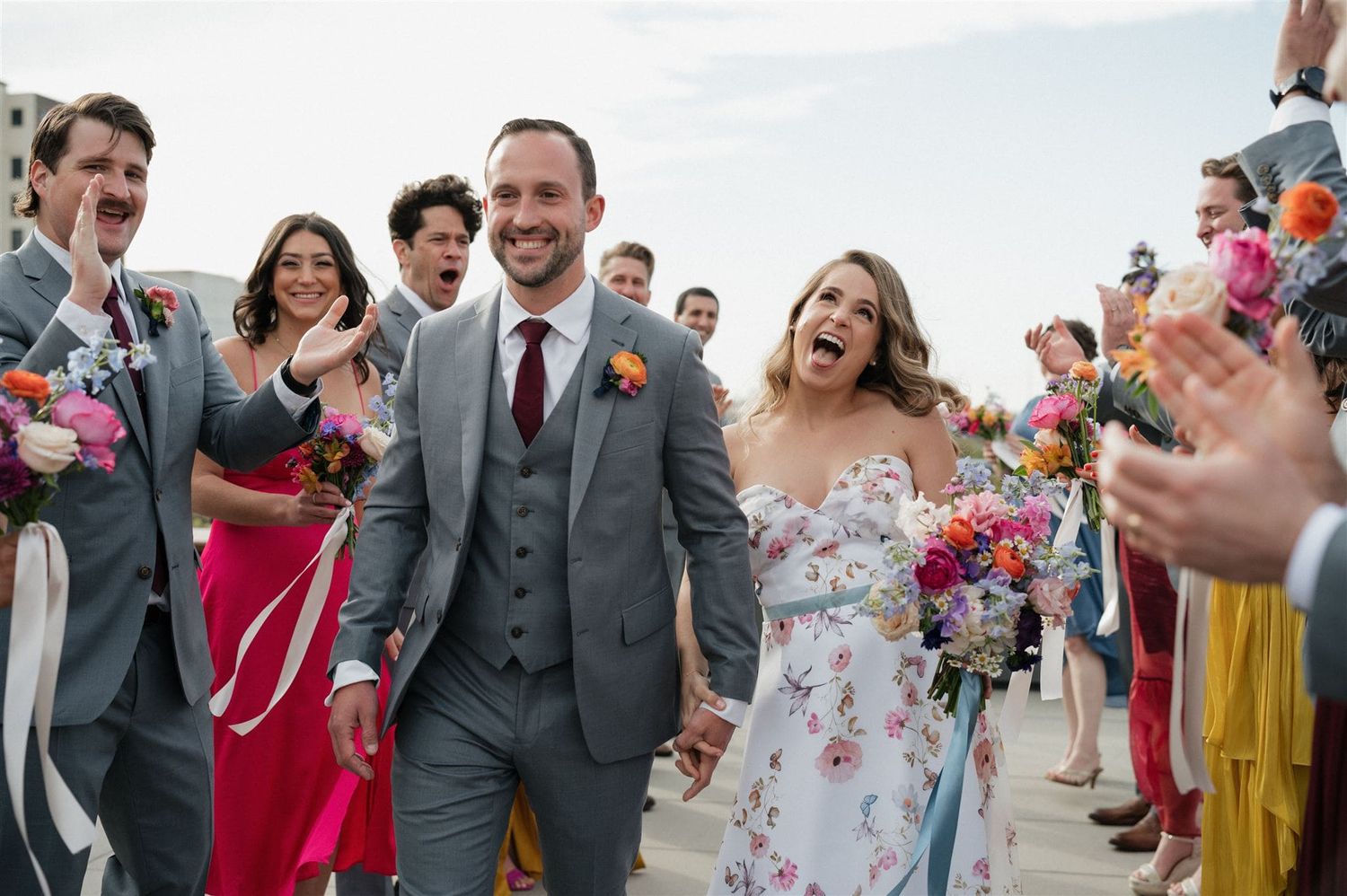 Wedding celebration on a sunny day as happy newlyweds walk through a cheering crowd of guests in colorful attire.