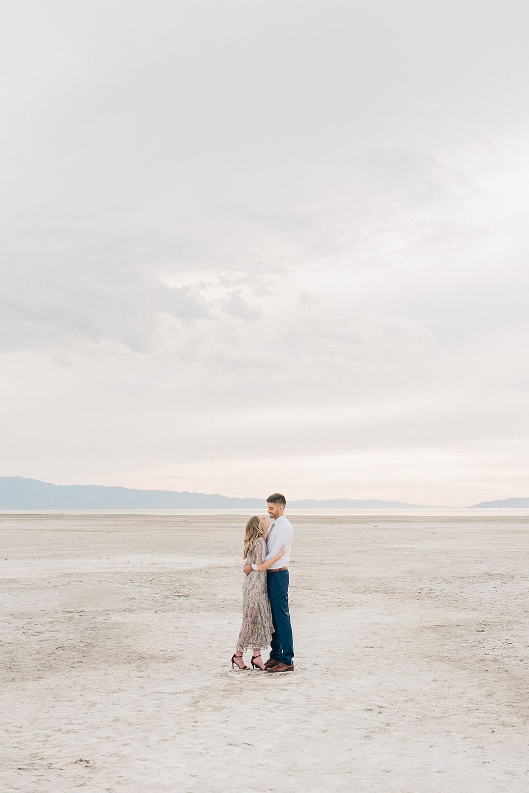Romantic desert engagement photo with couple standing on salt flats under cloudy sky.