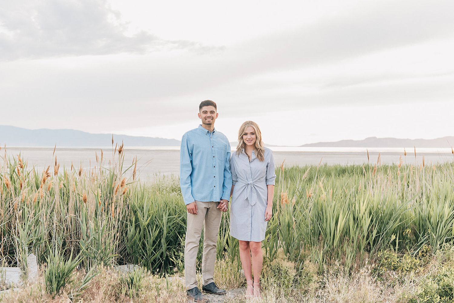A couple stands together in tall grass with mountains in the distance wearing coordinating light blue outfits.