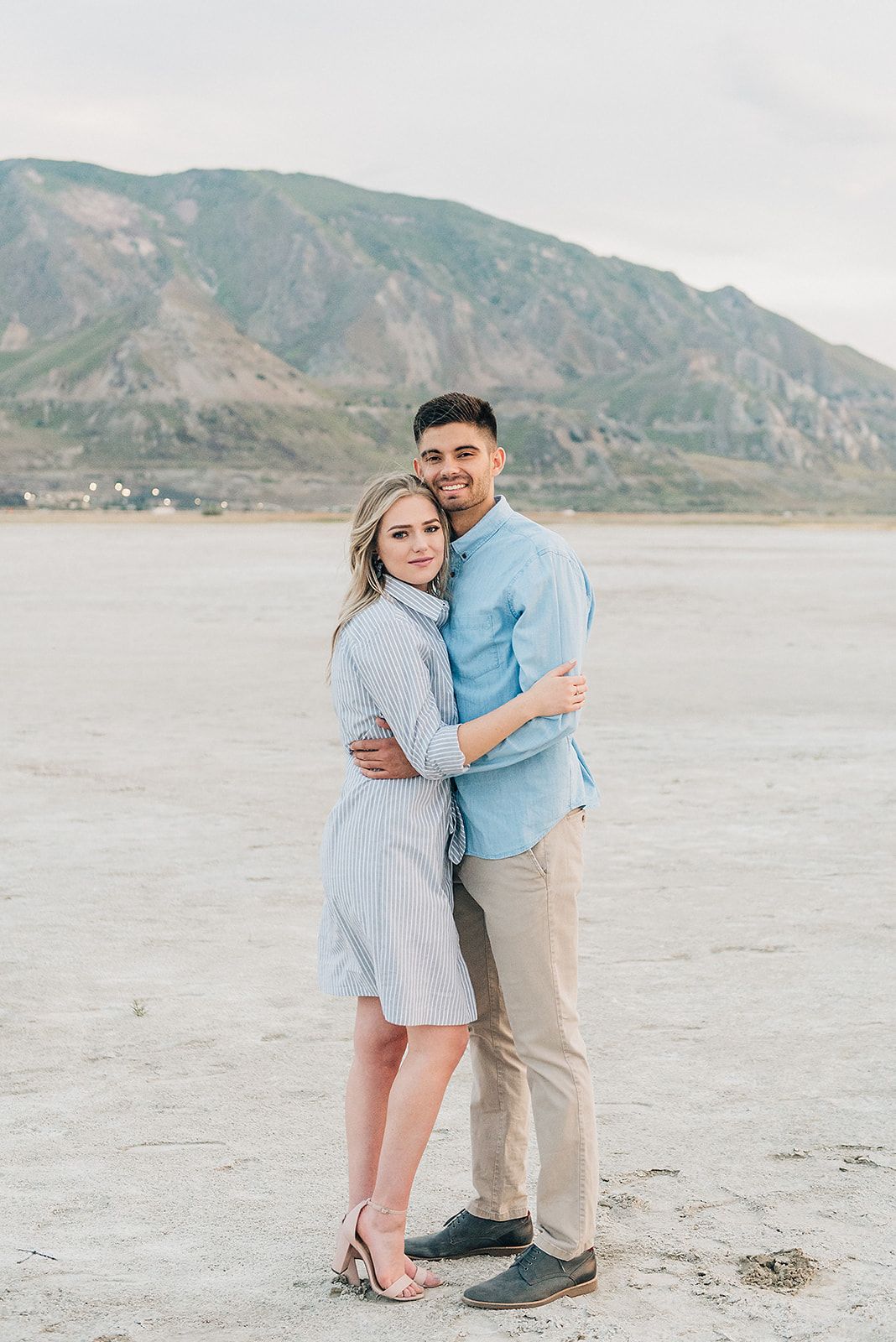 A couple embraces on a desert plain with dramatic mountain backdrop and soft lighting.