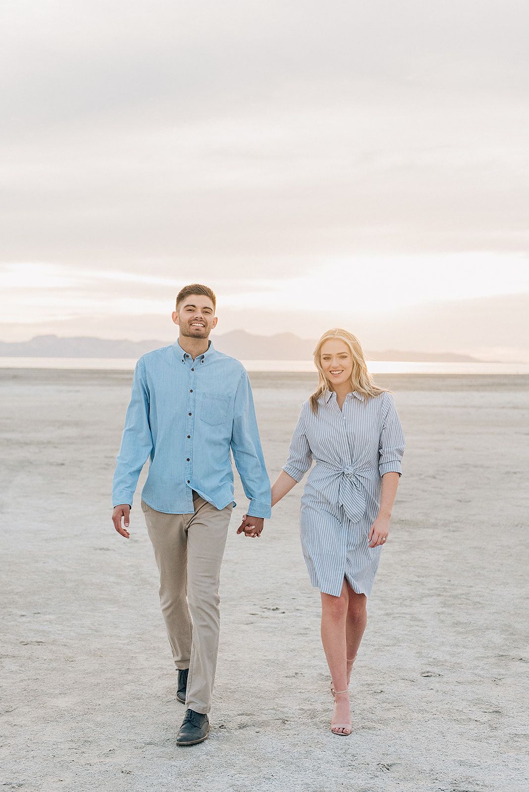 A couple walks hand in hand across a salt flat desert landscape at dusk.
