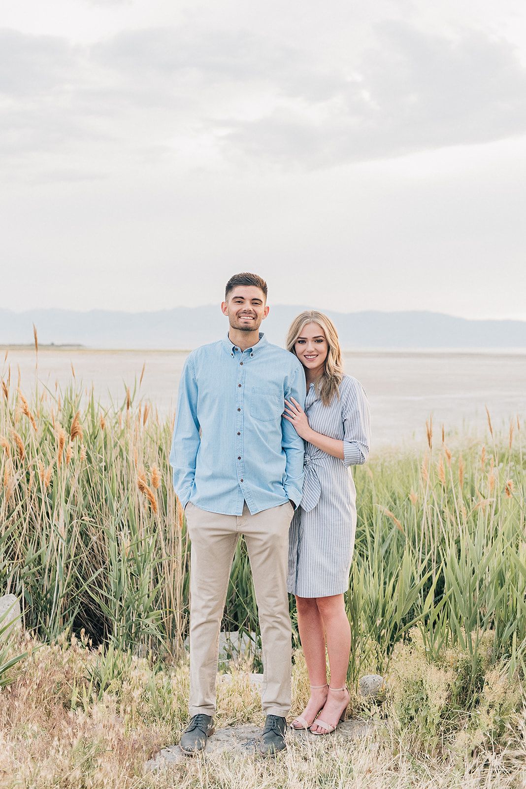 A couple poses in matching blue outfits among tall grasses with a lake view.