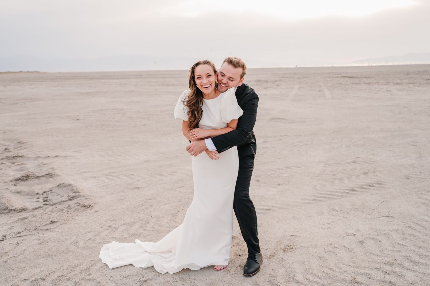 Newlywed couple embraces on sandy beach during sunset wedding photoshoot.
