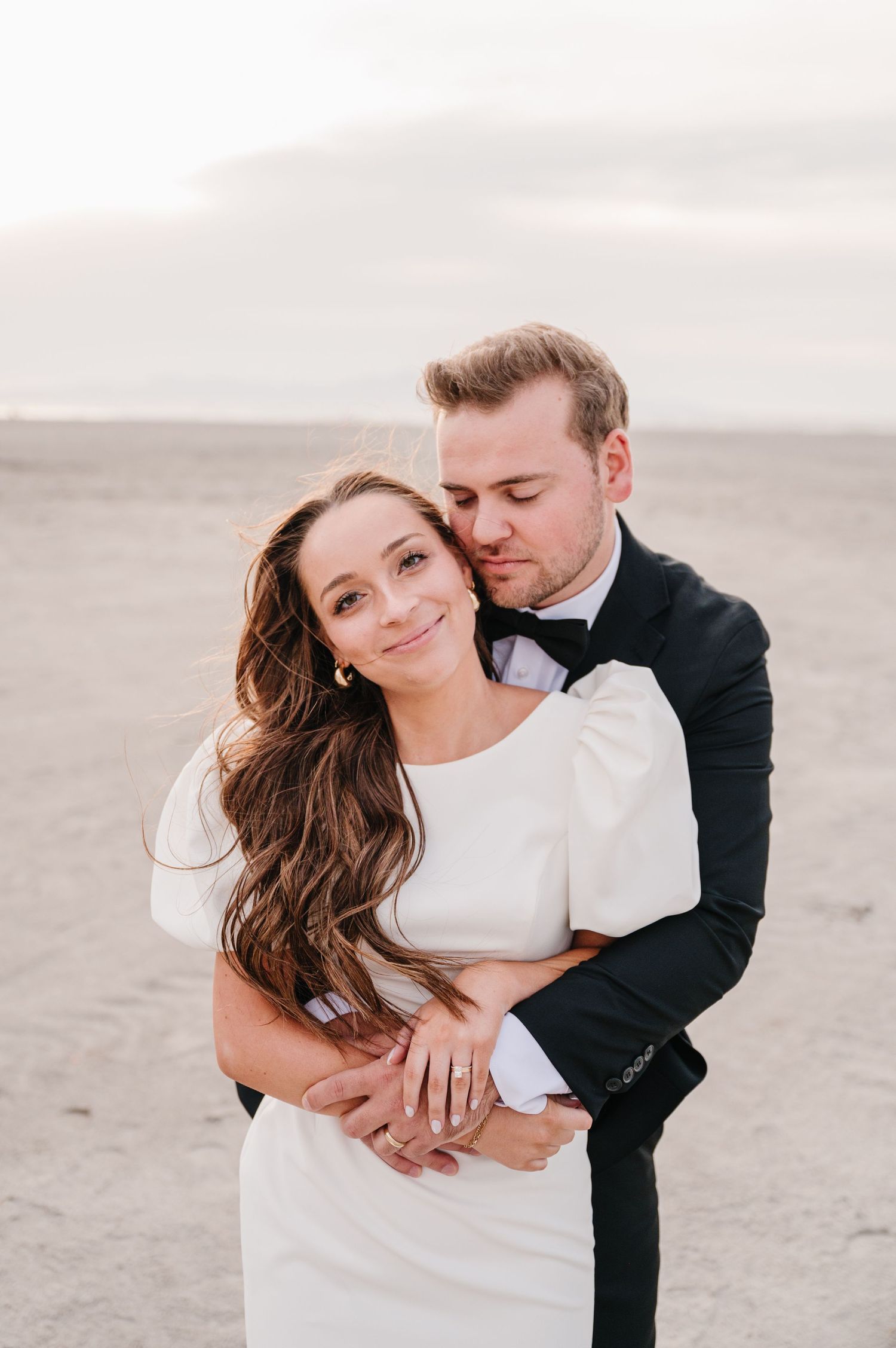 Romantic embrace on sandy beach at sunset in formal wedding attire.