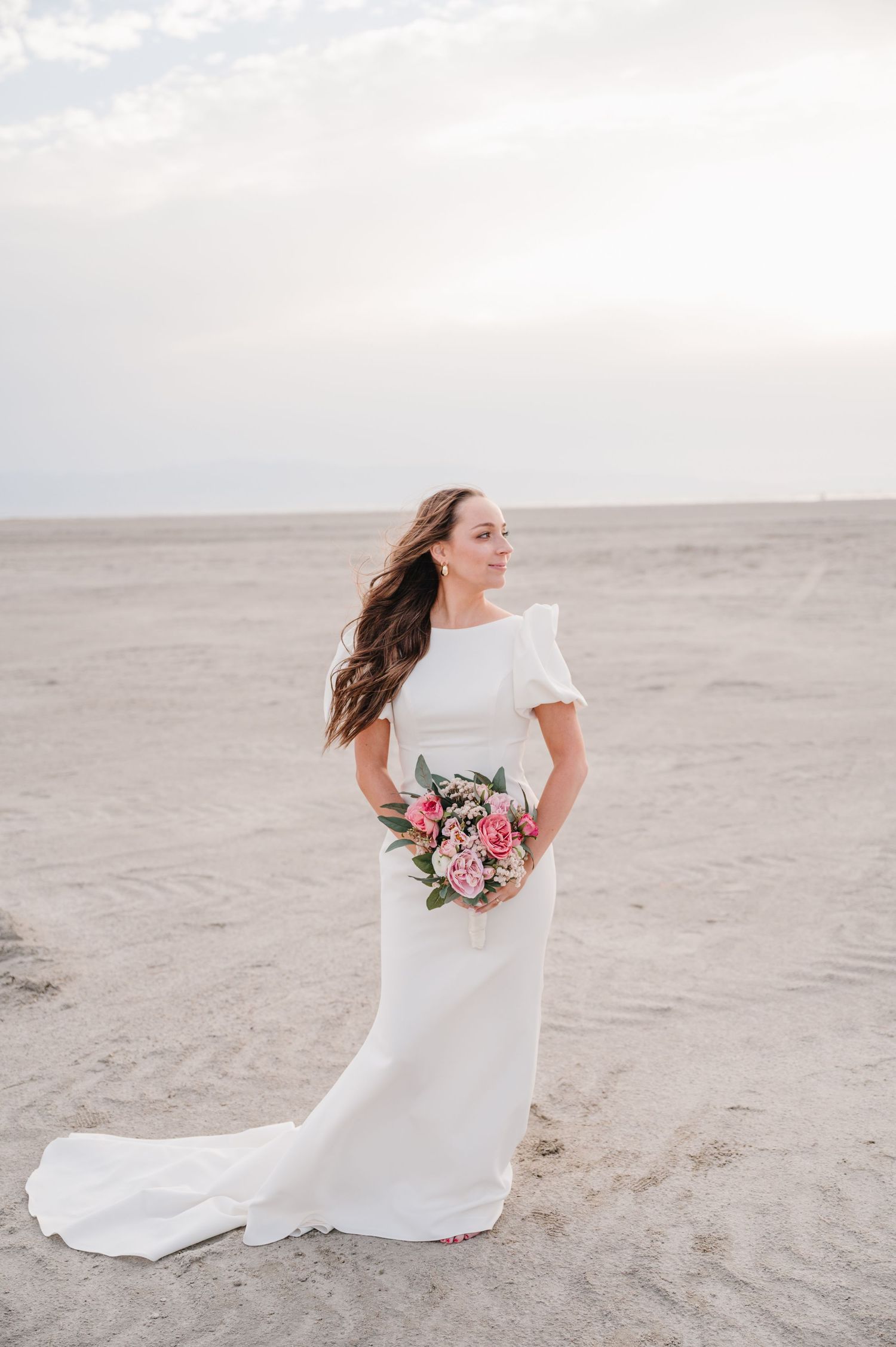 Bride in white dress holding bouquet on sandy beach with overcast sky.
