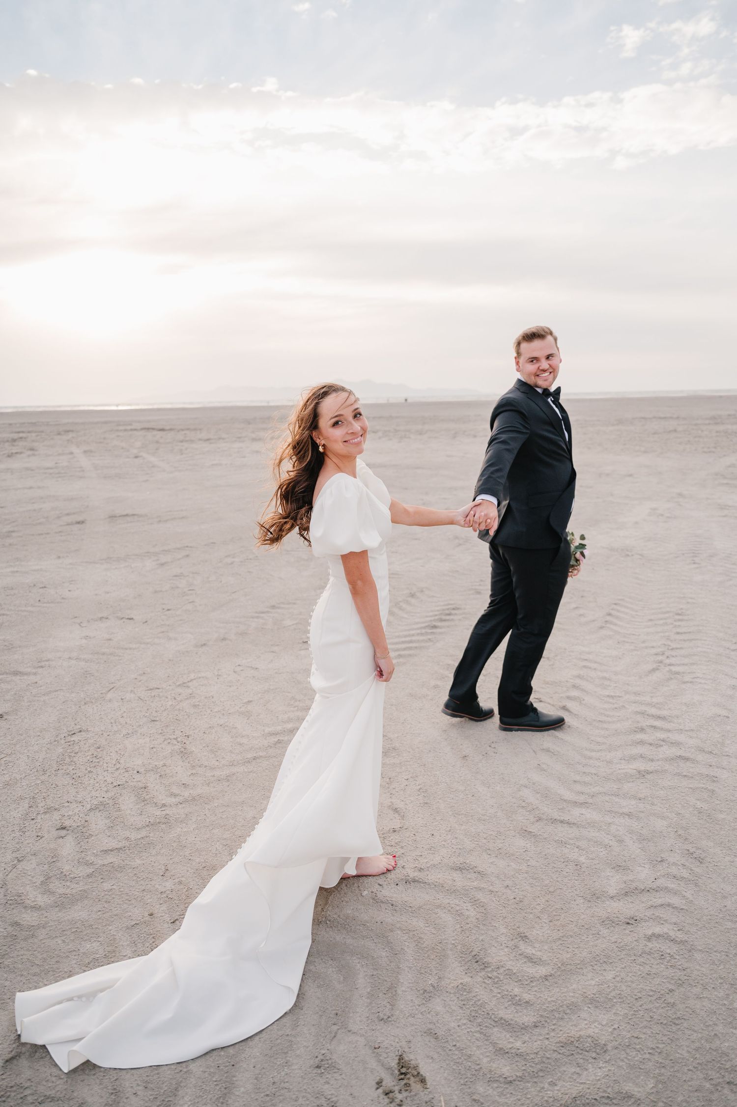 Couple in wedding attire playfully poses on sandy beach during sunset.