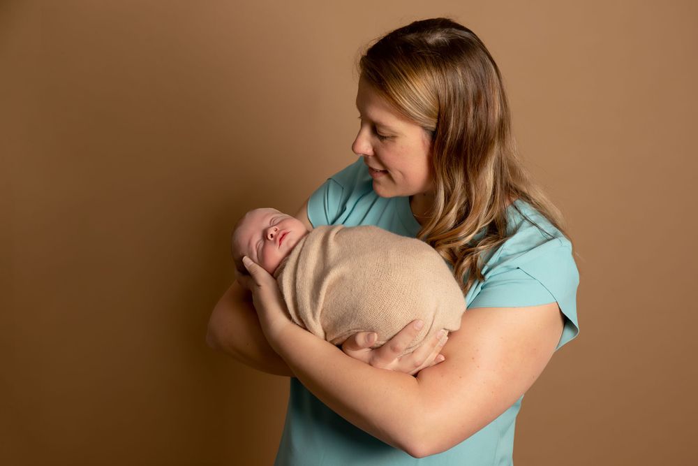 Mother in turquoise shirt cradles sleeping newborn wrapped in beige blanket against brown background.