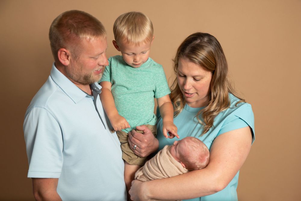 A young family poses together in coordinating blue outfits with their newborn baby and toddler against a tan backdrop.