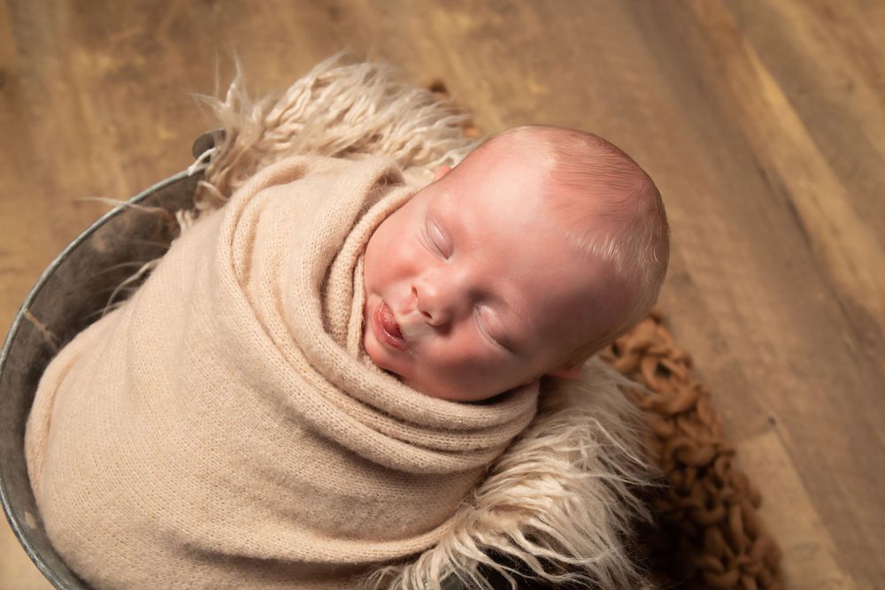 Newborn baby sleeping peacefully in metal bowl with fur lining and tan wrap against wooden floor.