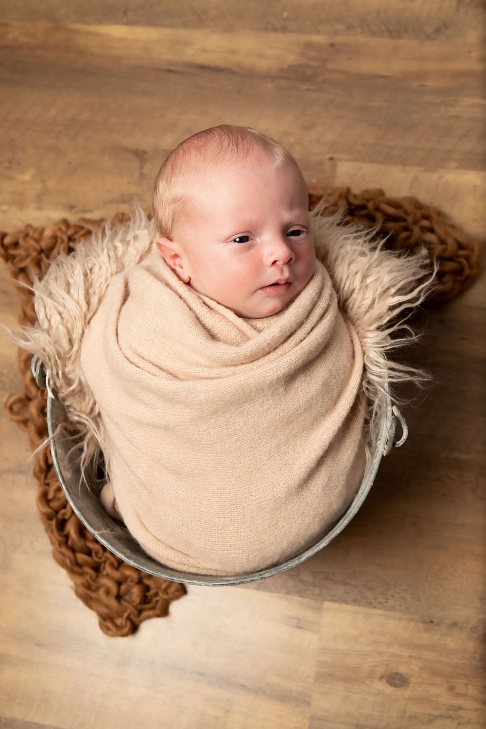 Peaceful newborn wrapped in tan swaddle rests in metal bowl with fur and wooden floor backdrop.