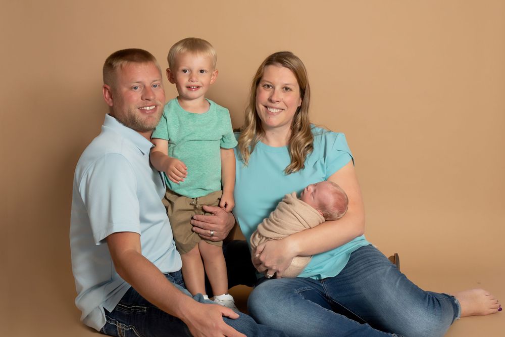 Casual family portrait with toddler and newborn sitting against warm brown background.