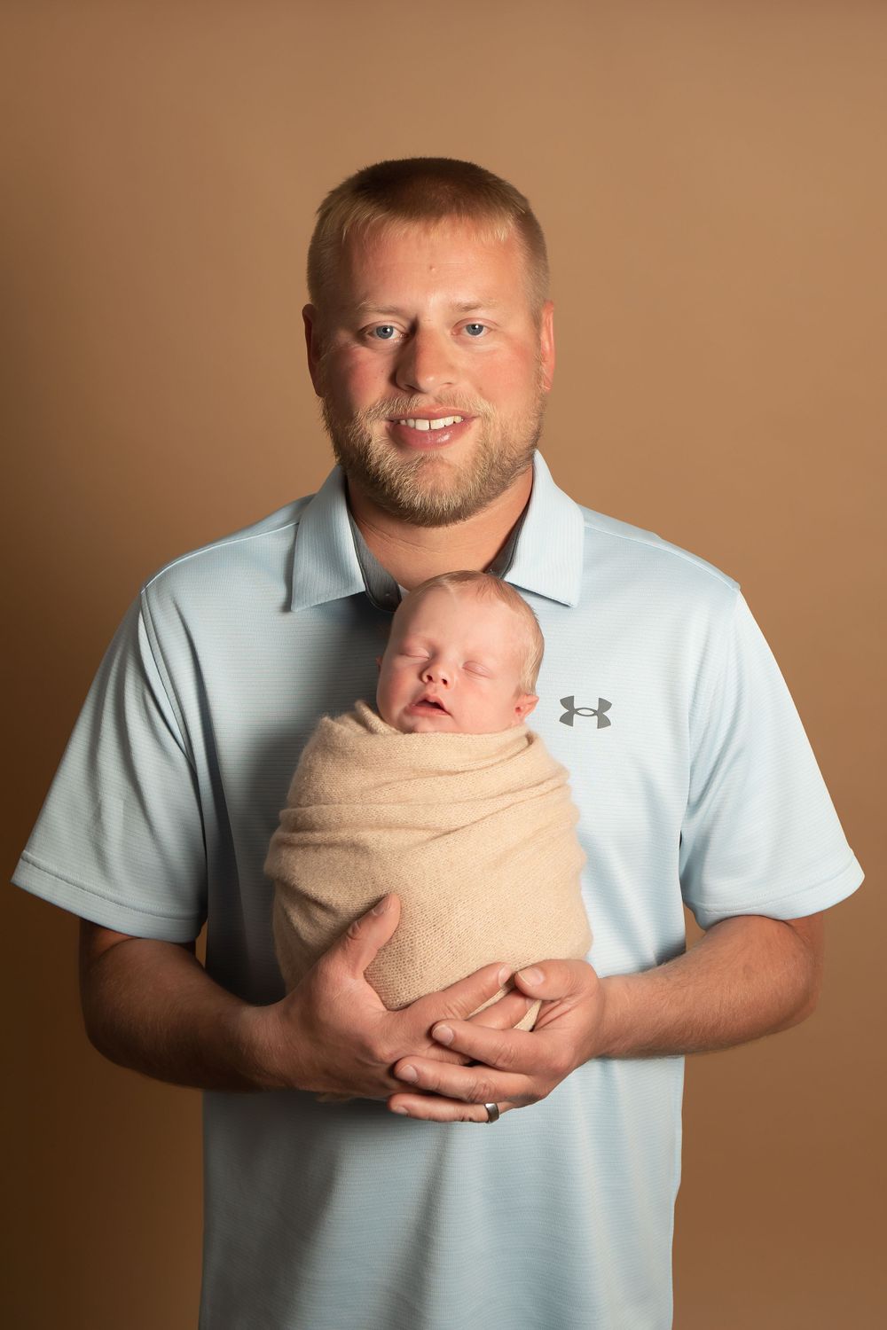 Father in light blue polo shirt holds sleeping newborn swaddled in tan wrap against brown backdrop.