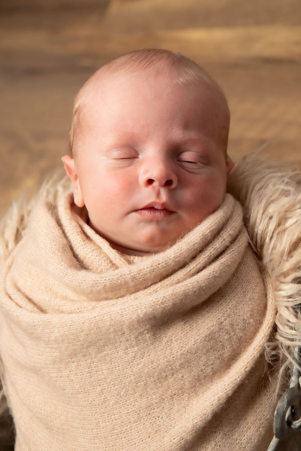 Sleeping newborn wrapped in soft beige swaddle photographed in close-up against wooden background.