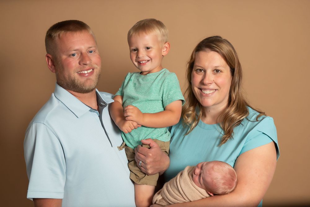 Family portrait with toddler in mint green shirt and newborn against brown background.