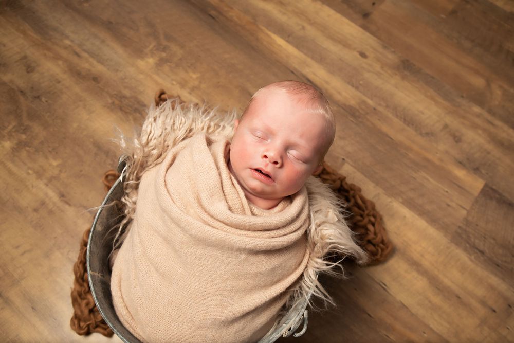 Overhead view of sleeping newborn in tan wrap posed in metal bowl on wooden floor.