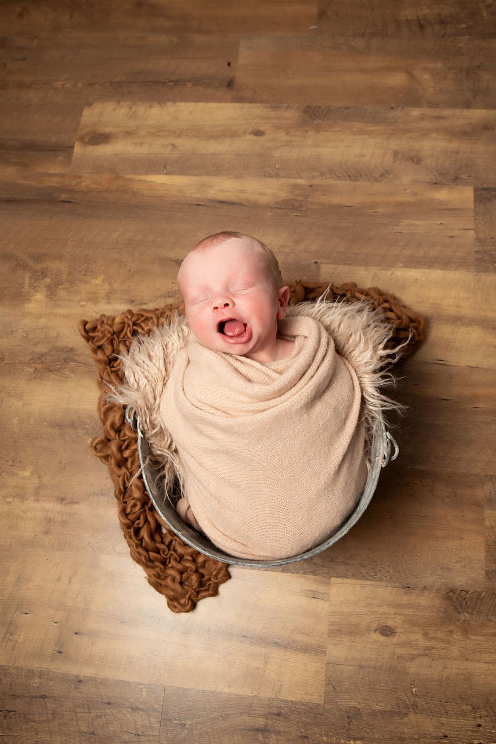 Newborn baby yawning while wrapped in beige blanket, posed in metal bowl with textured backdrop.