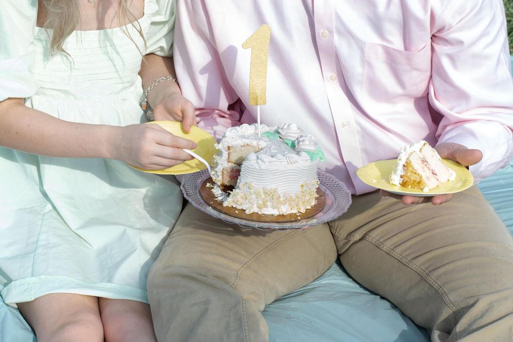A couple sharing slices of white frosted cake on a picnic blanket outdoors.