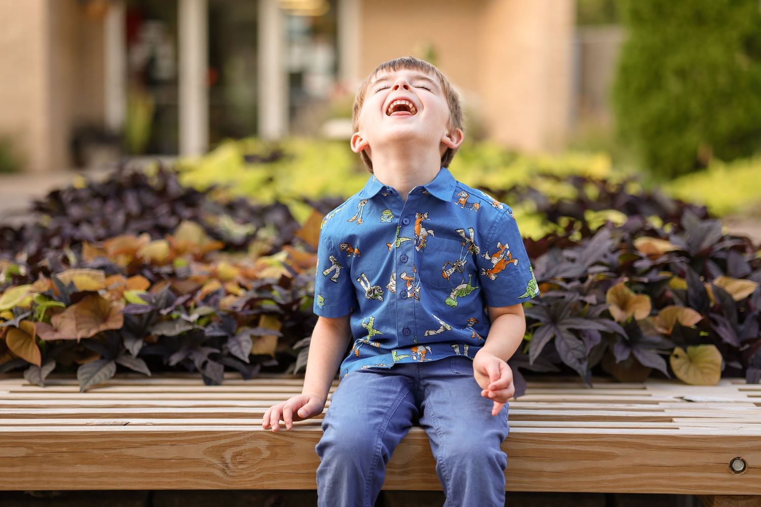 Young child laughs joyfully while sitting on wooden bench with landscaping in background.