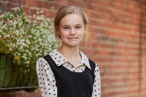 Young student in black and white polka dot dress stands in front of brick wall with blooming flowers.