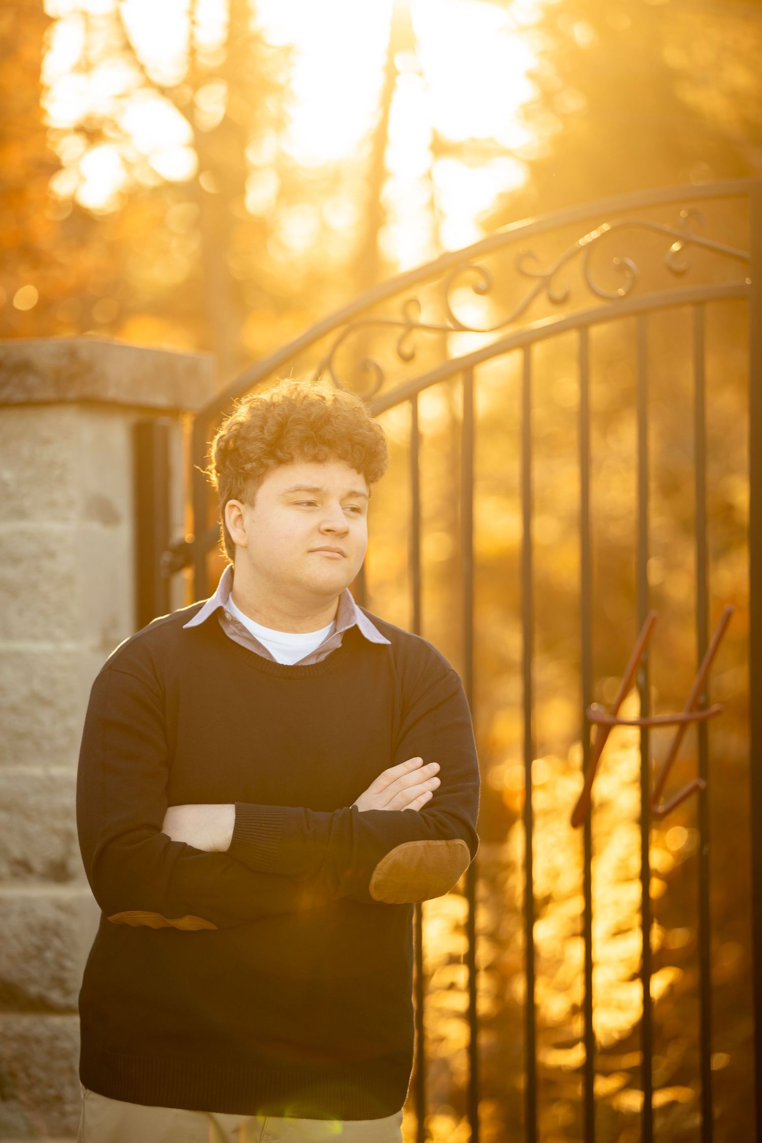 Figure in black sweater stands against ornate metal fence during golden sunset creating dramatic silhouette effect.