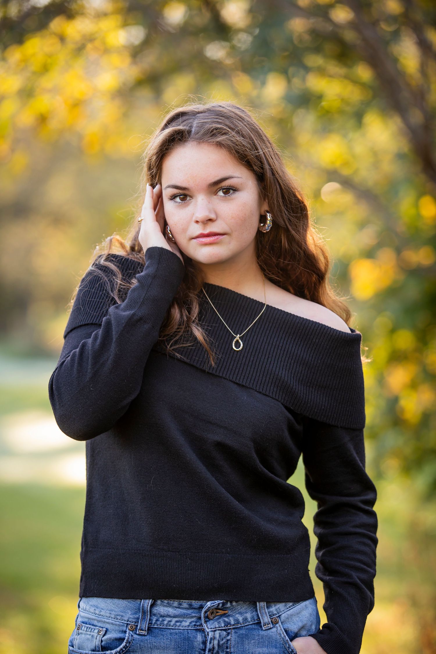 A model poses outdoors in a black sweater and jeans against a golden autumn background.
