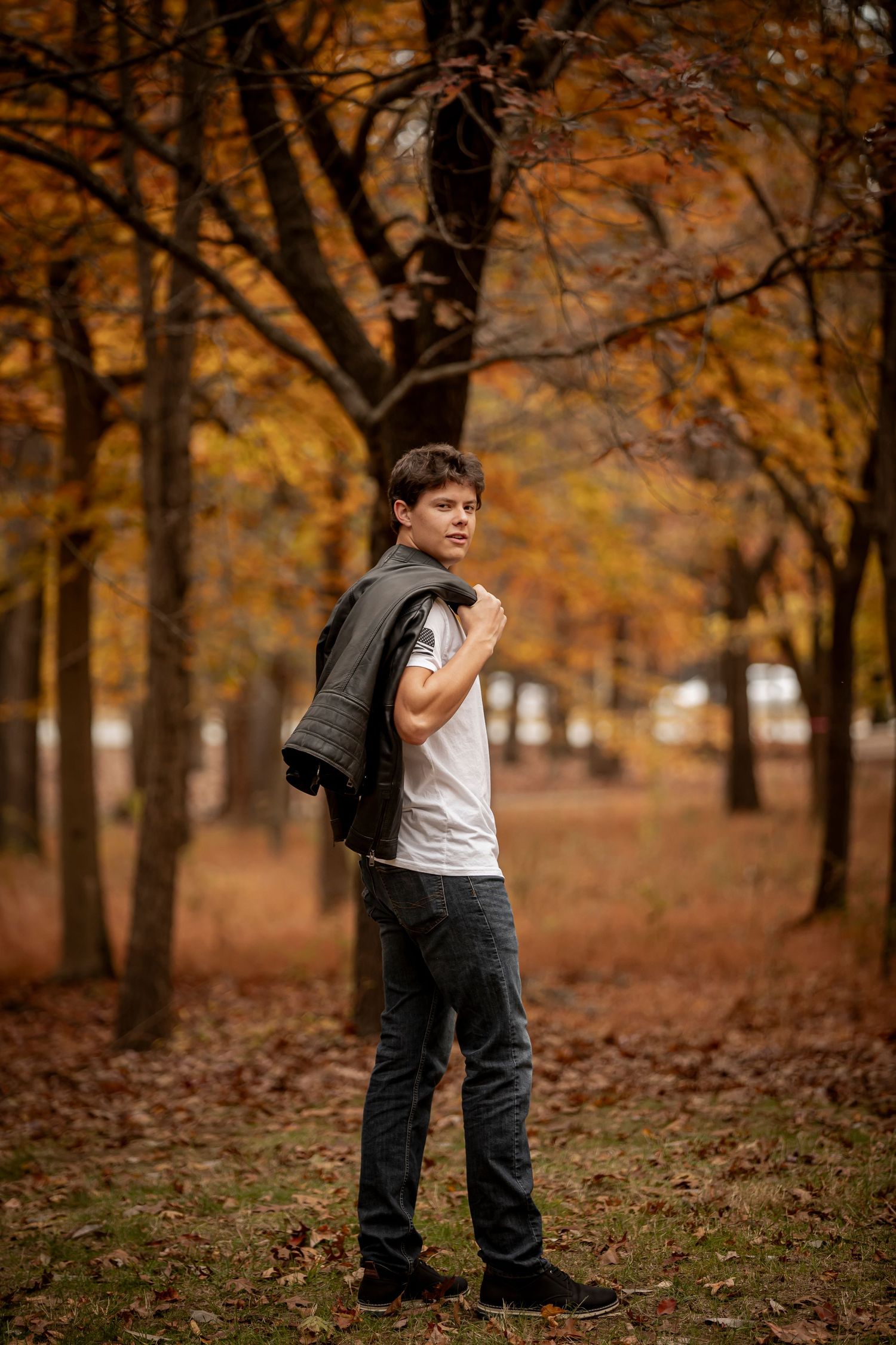 A child stands in a park filled with autumn trees and fallen orange leaves while wearing casual clothing.