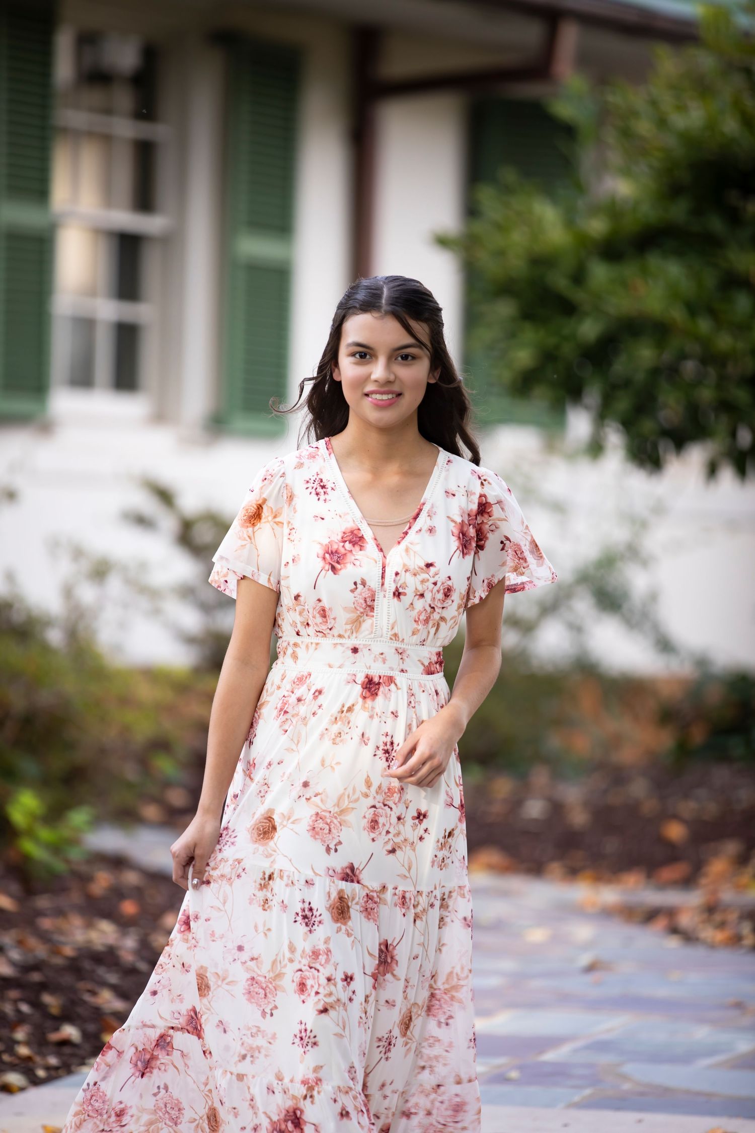 A person wearing a white and red floral print dress poses outdoors near a garden path.