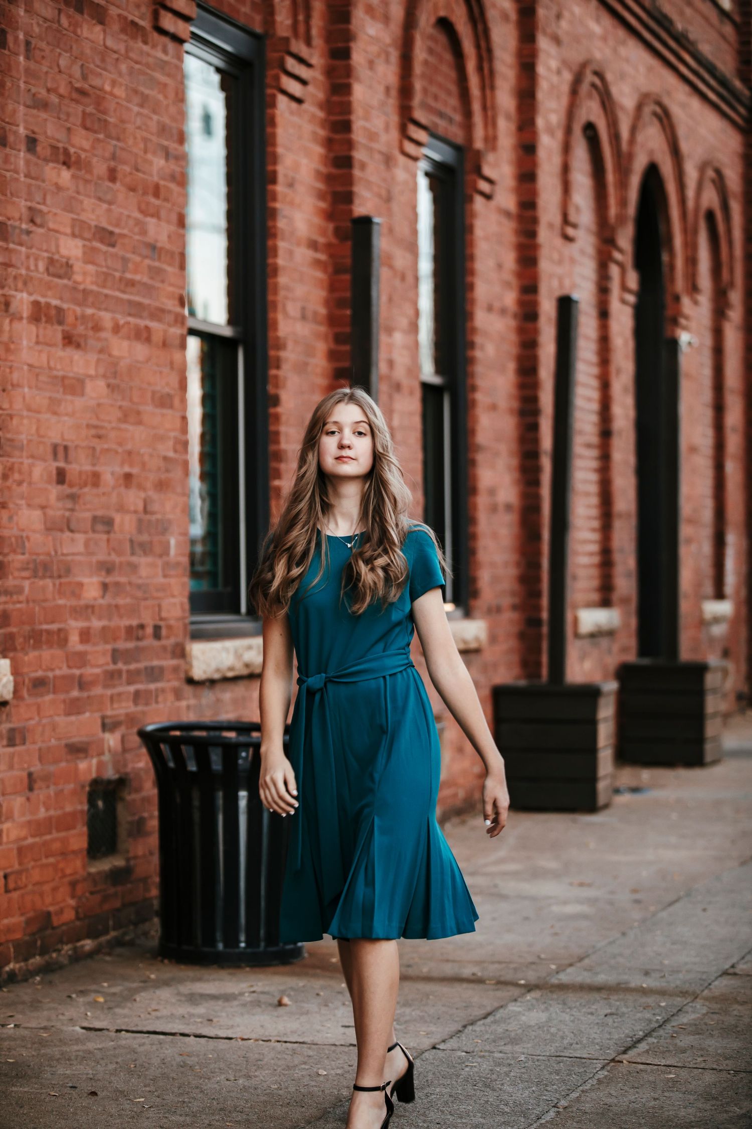 A child in a teal dress twirls and dances along a brick wall walkway with black shuttered windows.