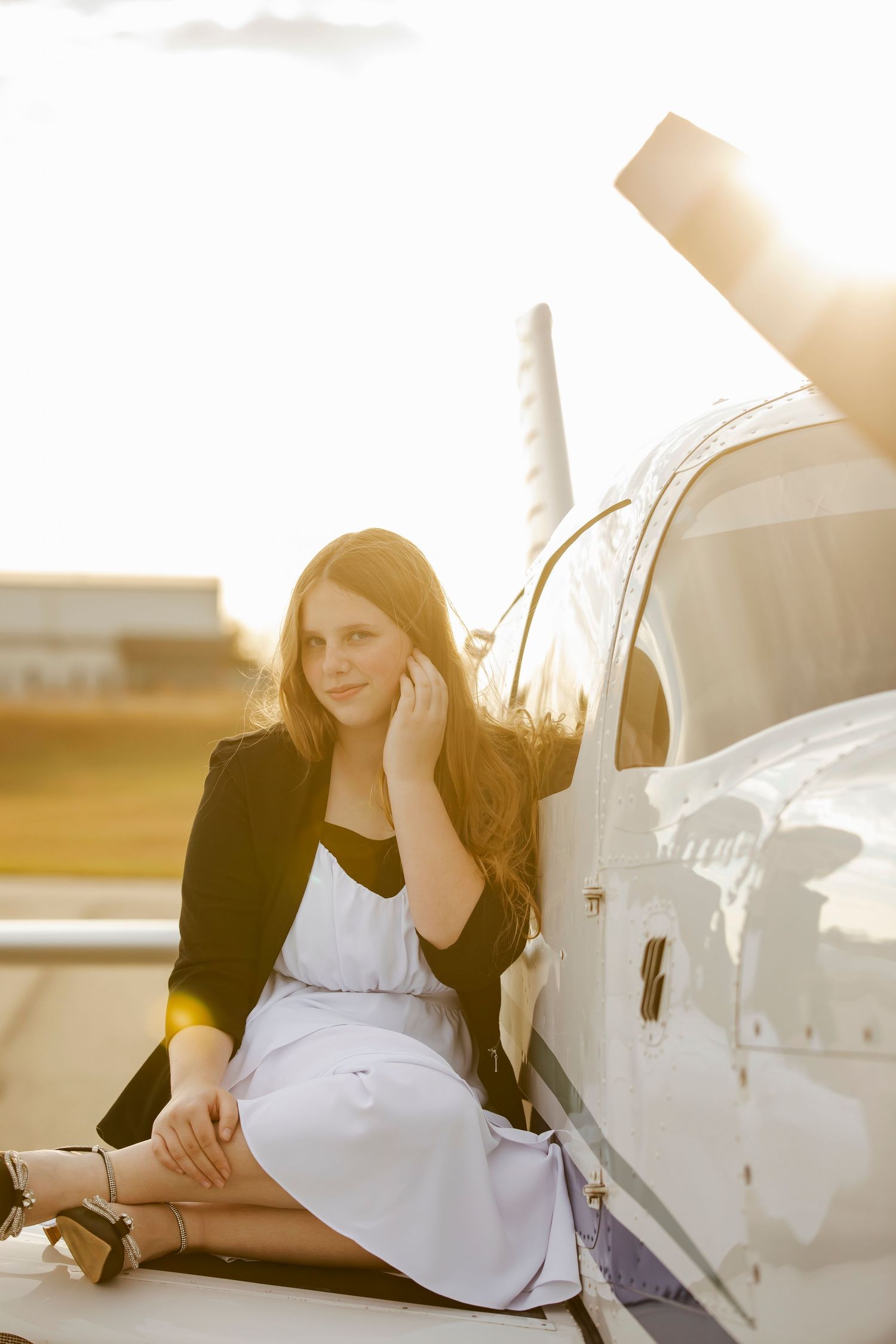 Woman in black and white outfit leaning against small airplane during golden hour sunset.