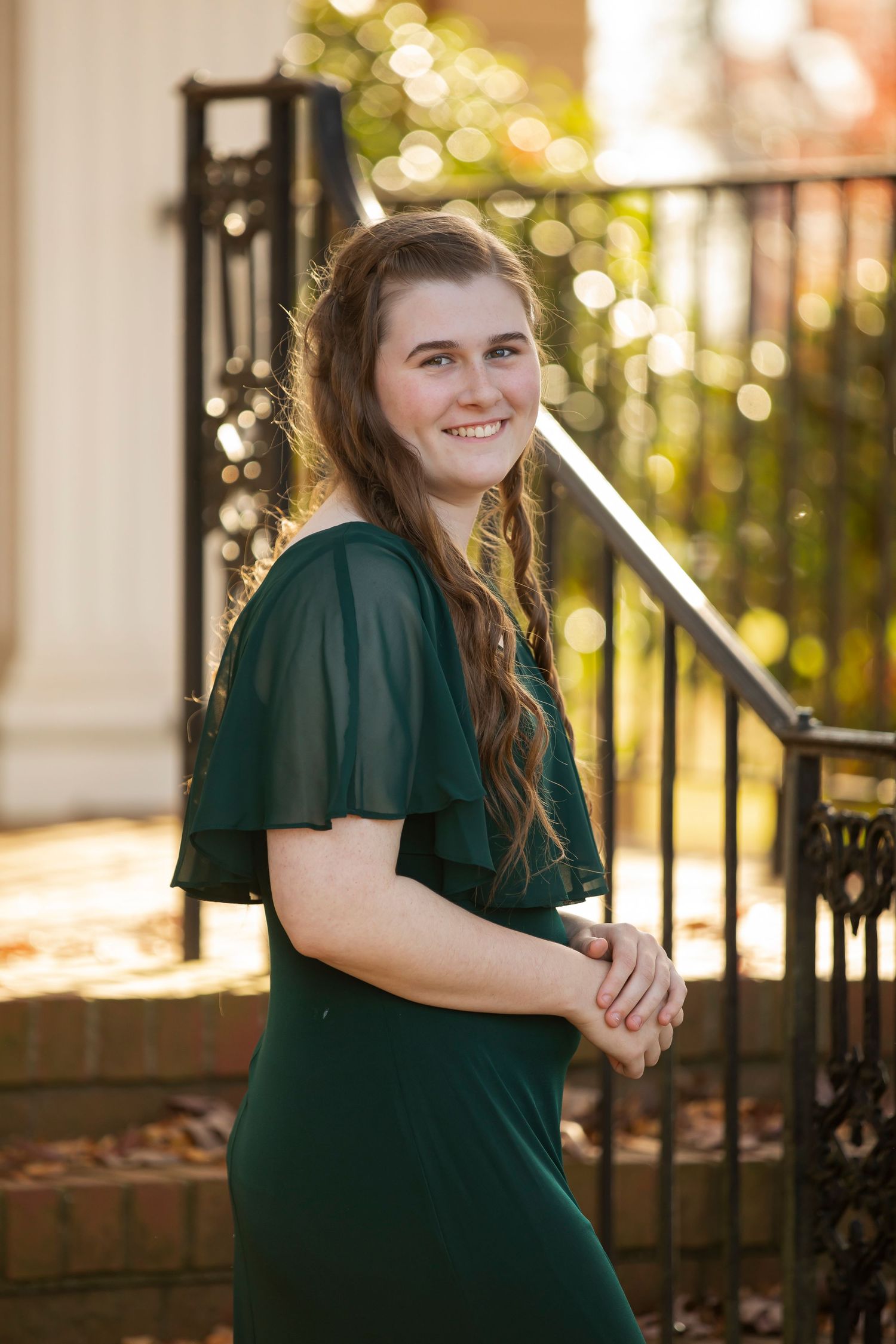 Graduate in dark green cap and gown poses outdoors with diploma in front of iron gate at sunset.