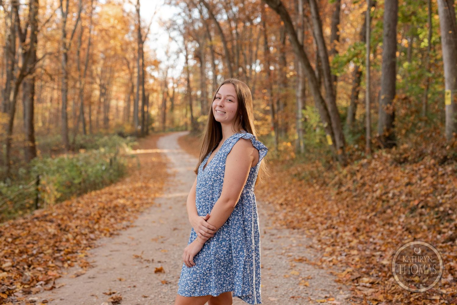A person in a blue floral dress poses on an autumn trail surrounded by golden fall foliage.