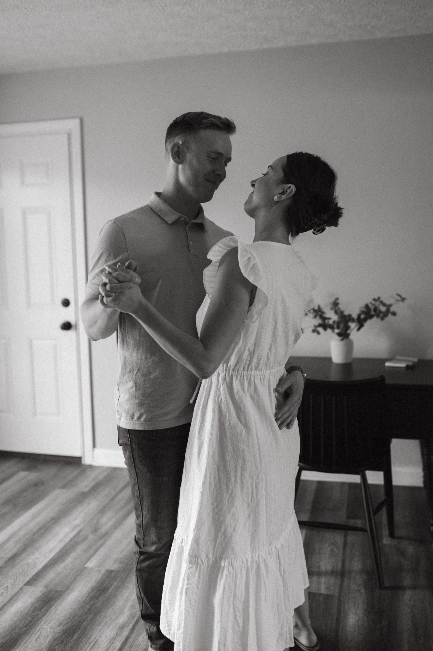 A black and white image shows a couple dancing and laughing together at home.