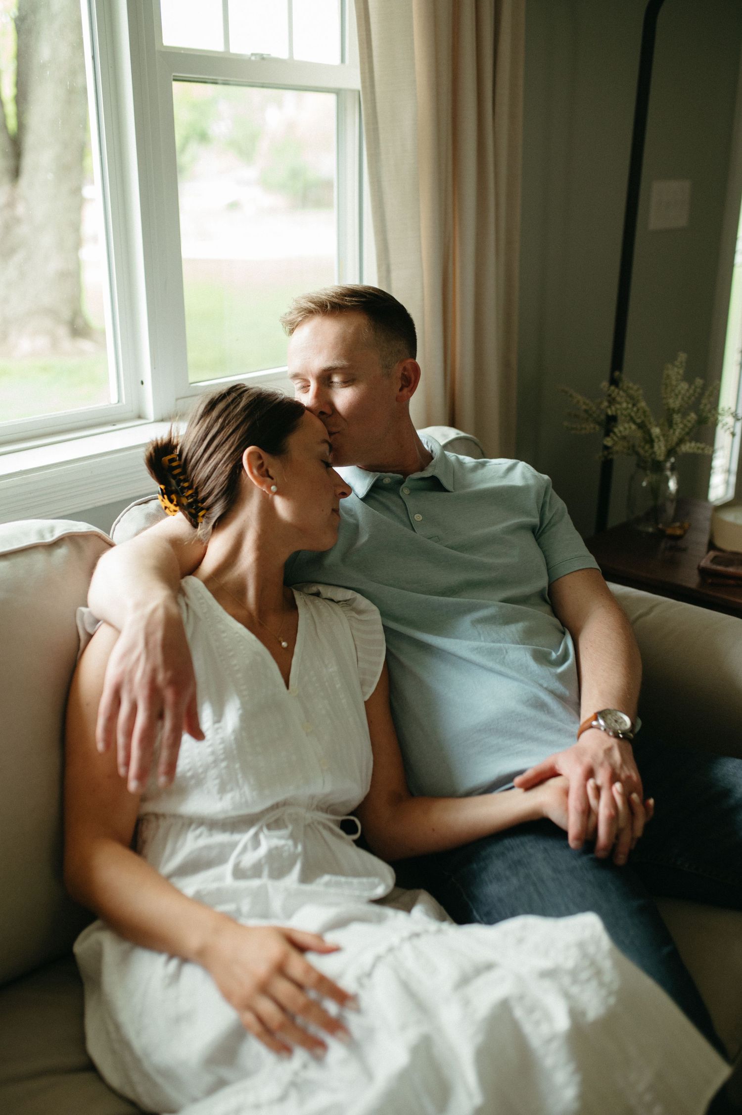 An intimate couples portrait by a bright window in casual attire on a cozy couch.