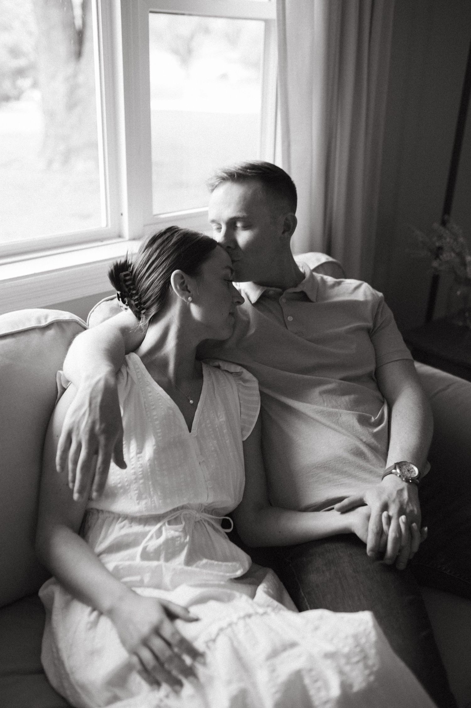 A tender black and white portrait of a couple embracing on a couch by a window.