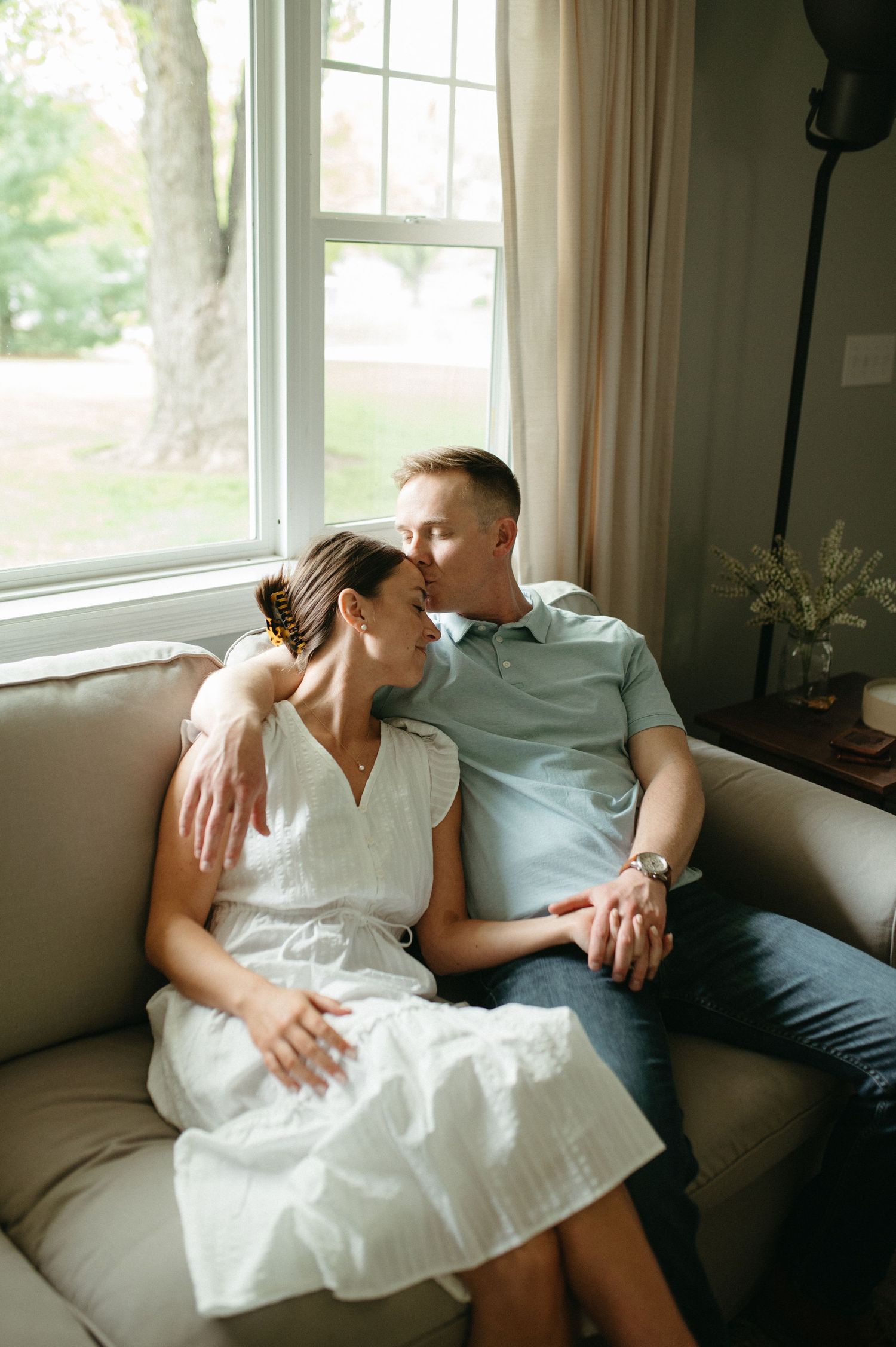 A couple relaxes together on a gray couch near a bright window with natural lighting.